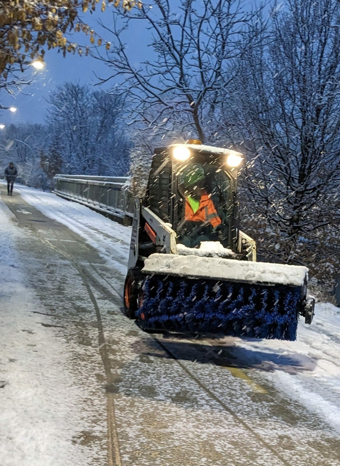 Worker with bright yellow safety vest drives a small snow plow vehicle on a snowy walkway.