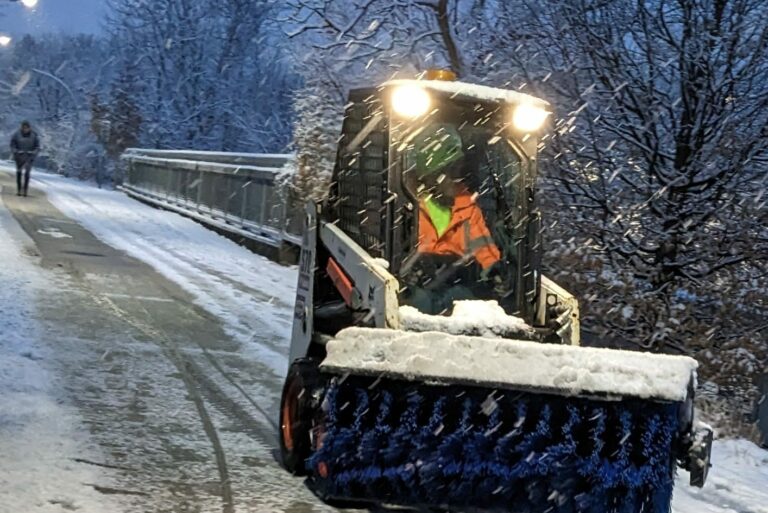 Worker with bright yellow safety vest drives a small snow plow vehicle on a snowy walkway.