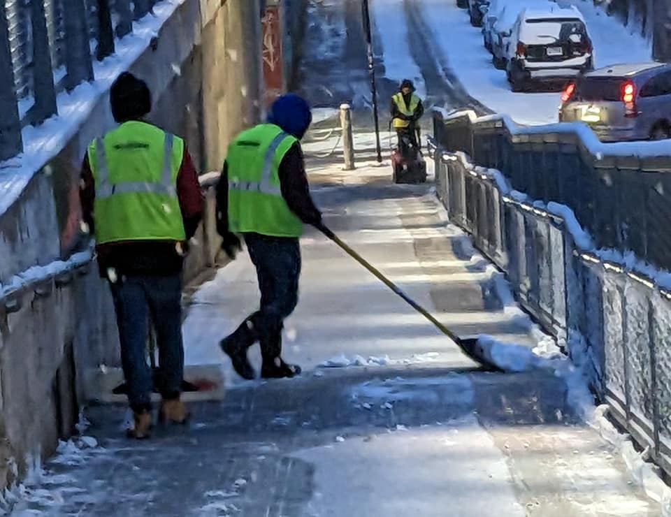Workers wearing bright yellow safety vests shovel a snowy ramp walkway