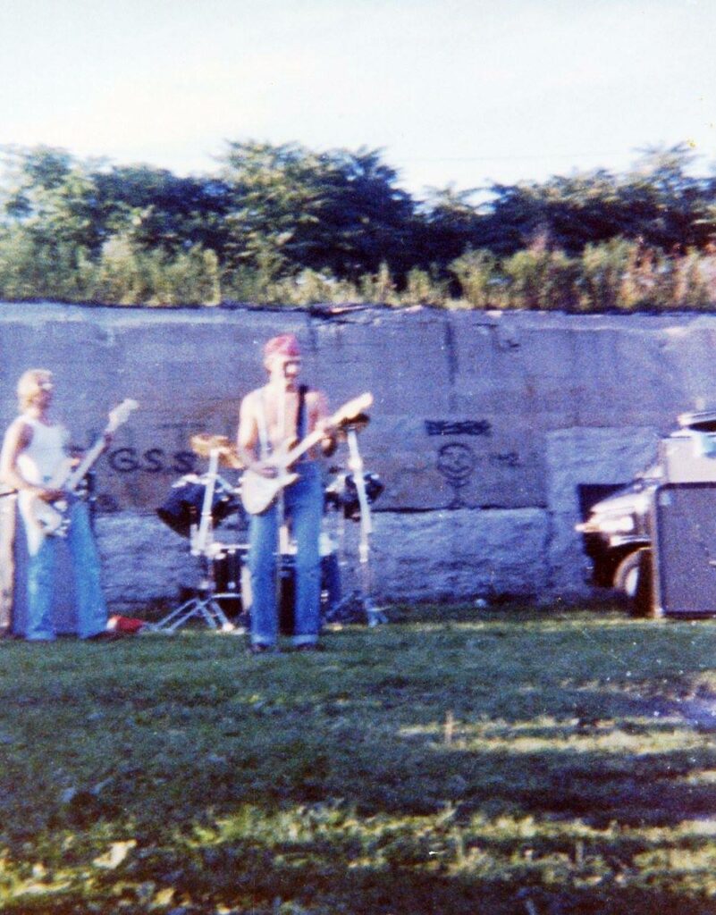 Grainy photo of a rock band playing outside on the grass in front of a wall with graffiti on it.