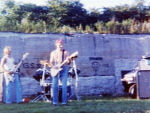 Grainy photo of a rock band playing outside on the grass in front of a wall with graffiti on it.