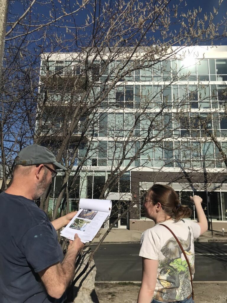 Two people facing a tree and building, one taking notes.