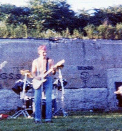 Grainy photo of a rock band playing outside on the grass in front of a wall with graffiti on it.